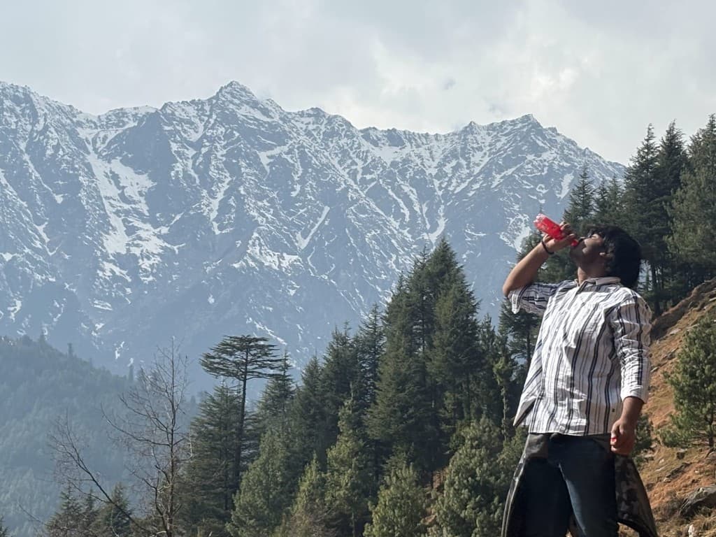 Vinayak on a mountain slope, snow peaks behind, red bottle in hand, very hireable energy.